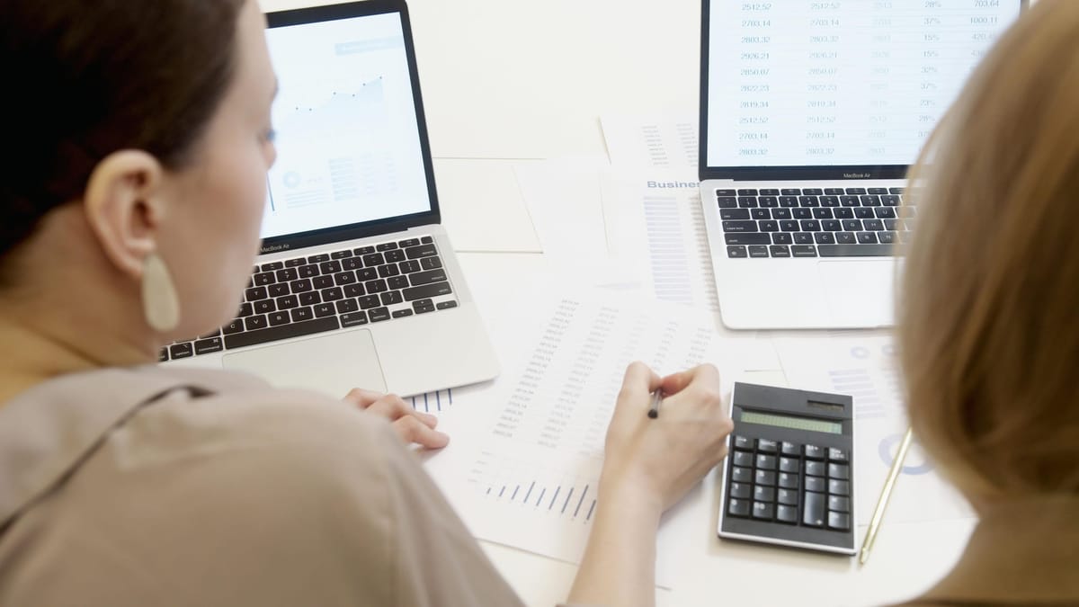 Two professionals analyze inventory and sales data using laptops, printouts, and a calculator during a business meeting.