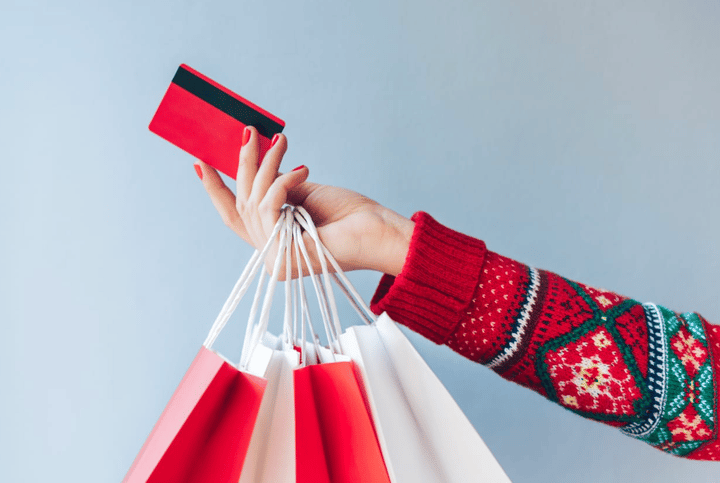 La mano de una mujer con un suéter navideño sosteniendo muchas bolsas de compras festivas y una tarjeta de crédito roja.