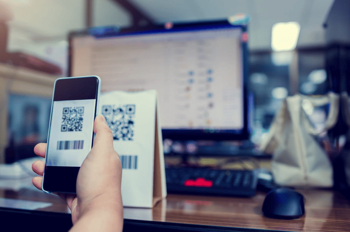 Una persona sosteniendo un teléfono inteligente para escanear un código QR con una pantalla de computadora borrosa al fondo