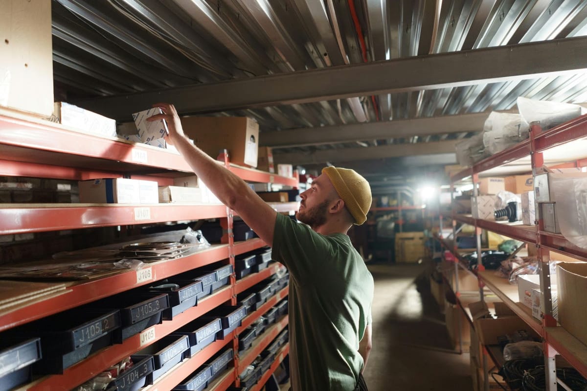 A warehouse worker in a green shirt and yellow beanie reaches up to retrieve a box from a high shelf in a warehouse with red metal shelving units.