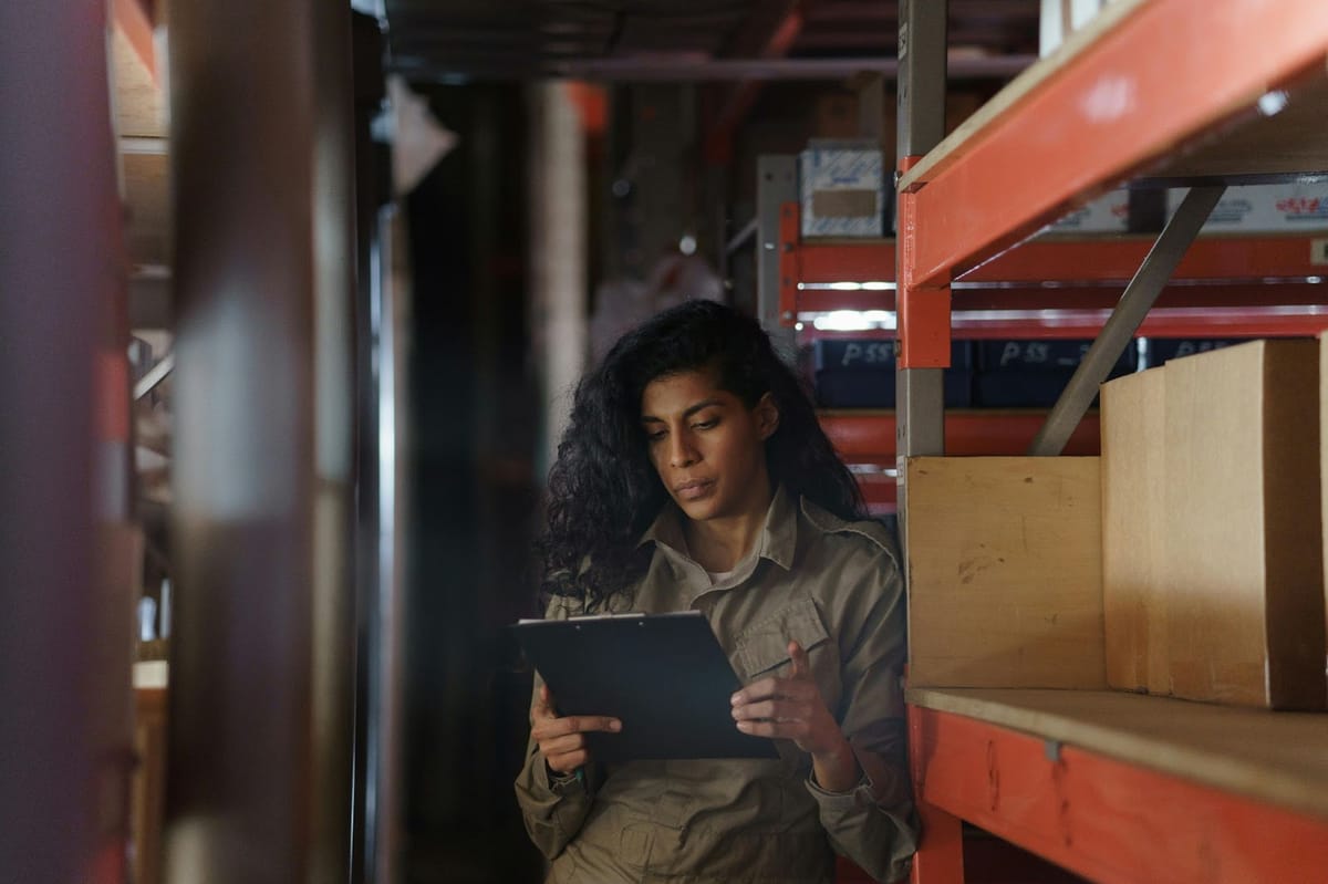 A warehouse worker in a khaki uniform reviews information on a tablet while leaning on a red metal shelving unit stocked with cardboard boxes.