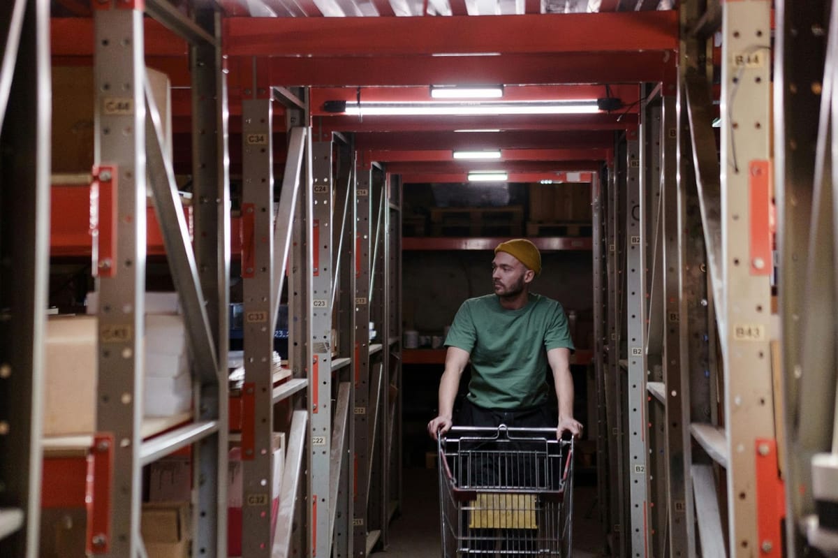 A warehouse worker pushes a wire shopping cart down a narrow aisle between tall red metal shelving units with location labels.