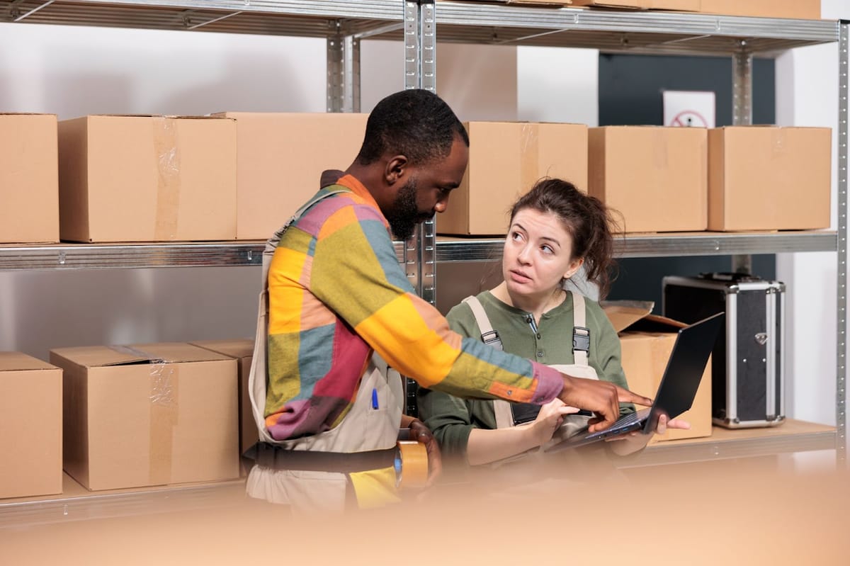 Two warehouse workers looking at a laptop together while surrounded by cardboard boxes on metal shelves.