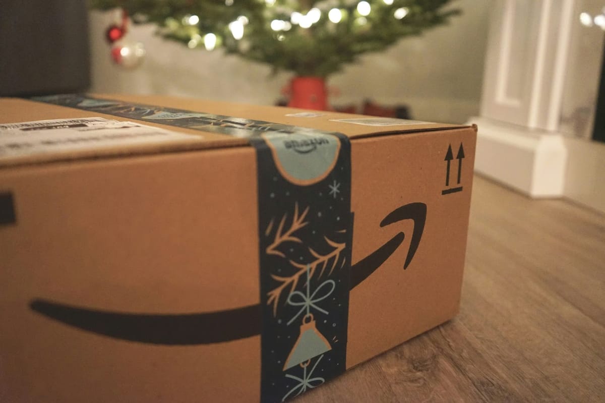 A festive Amazon package with holiday-themed tape placed on a wooden floor, with a decorated Christmas tree blurred in the background.