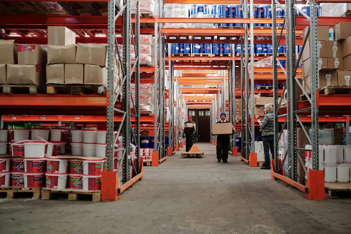 A wide view of a warehouse aisle with tall metal shelving units loaded with pallets of boxes and containers, with workers in the distance handling inventory items.