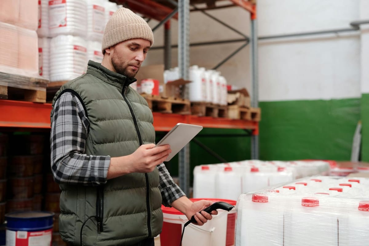 A person examines a tablet while holding a scanner next to pallets of white containers with red lids in a warehouse.