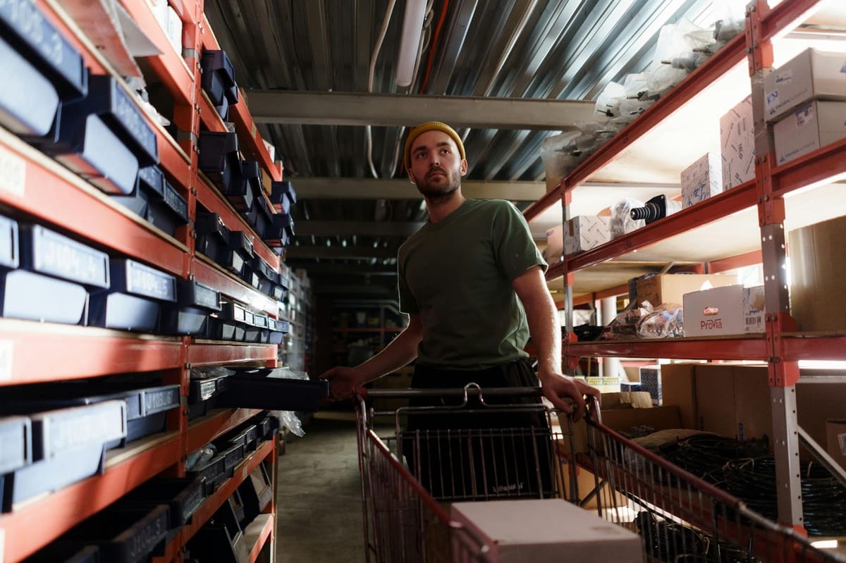 A person wearing a yellow beanie and green t-shirt pulls a wire cart along warehouse shelving units filled with stacked black storage bins and organized inventory items