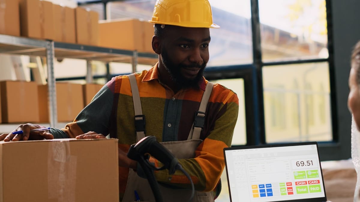  A person holding a carton and standing next to a barcode scanner and computer screen, with stacked boxes on a shelf in the background.