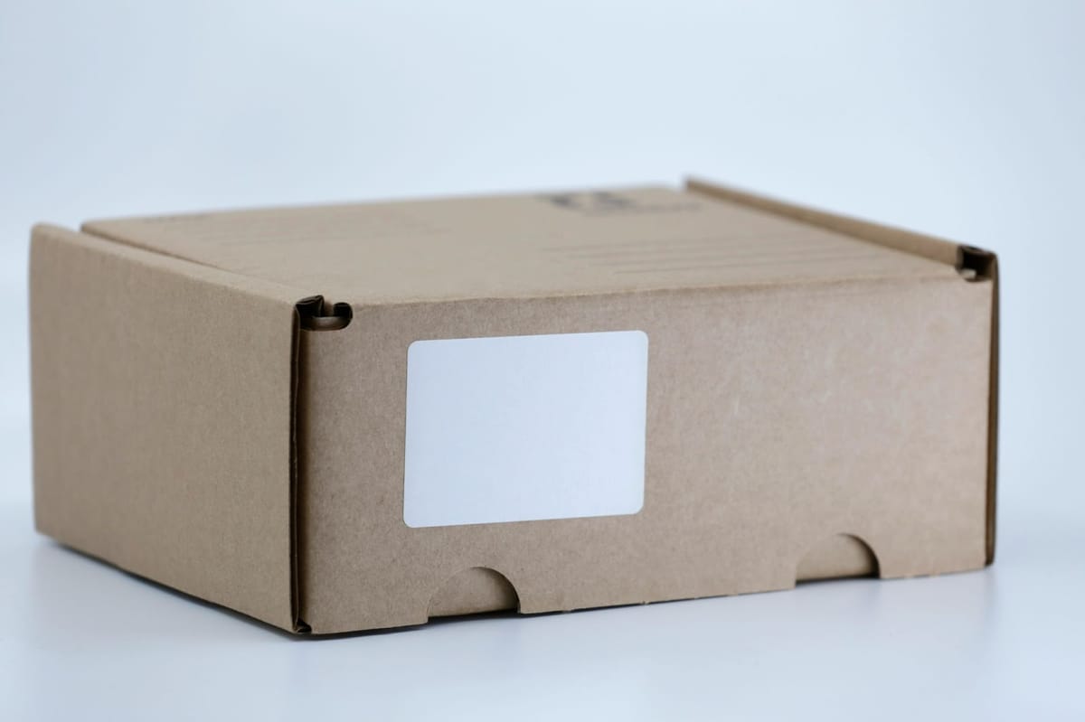 A closed, plain brown cardboard box with a blank white label on a white surface.