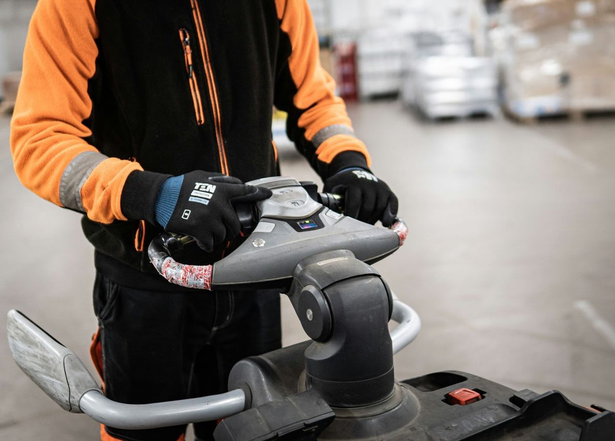 A worker in an orange and black uniform operating a pallet jack in a warehouse, holding the handle with gloved hands for precise control.