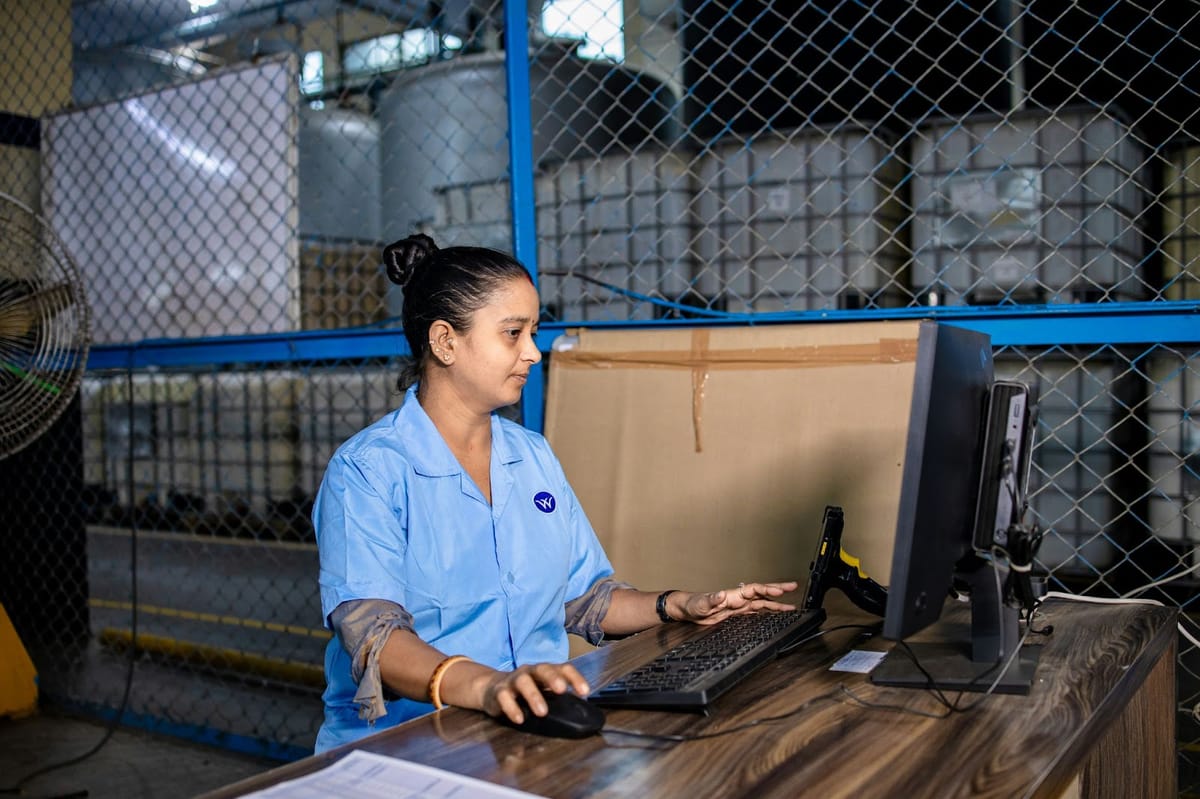 A person in a light blue uniform shirt sits at a computer workstation in a warehouse, with wire fencing and storage containers in the background.