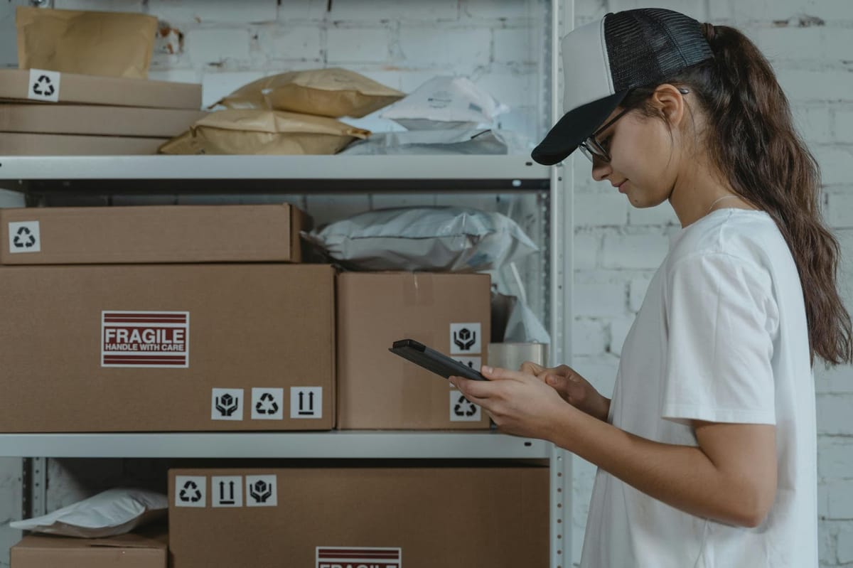A warehouse worker wearing glasses and a black cap uses a mobile device while standing next to shelves stocked with cardboard boxes marked with shipping symbols.