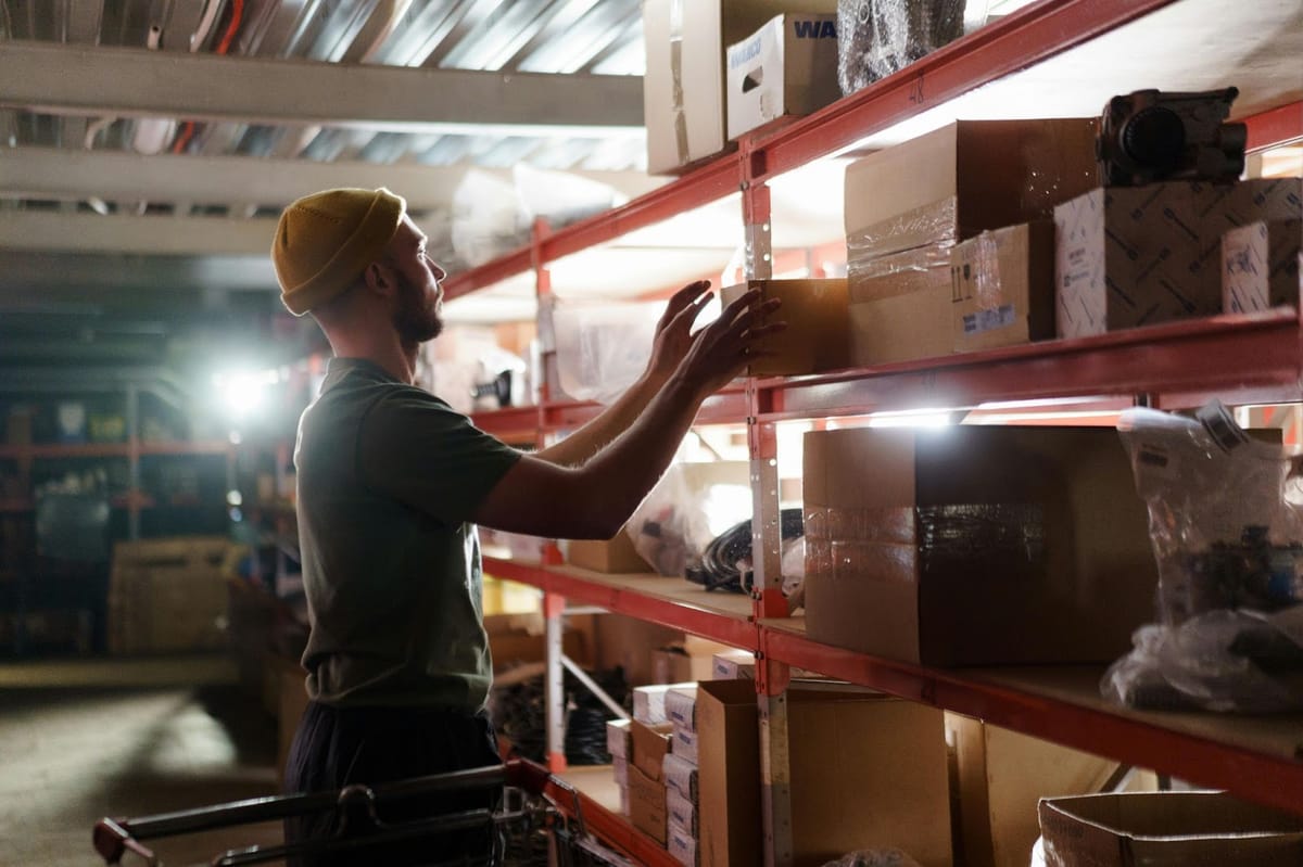 A person wearing a yellow beanie and green t-shirt reaches for boxed items on red metal shelving units in a warehouse storage area filled with packaged inventory.