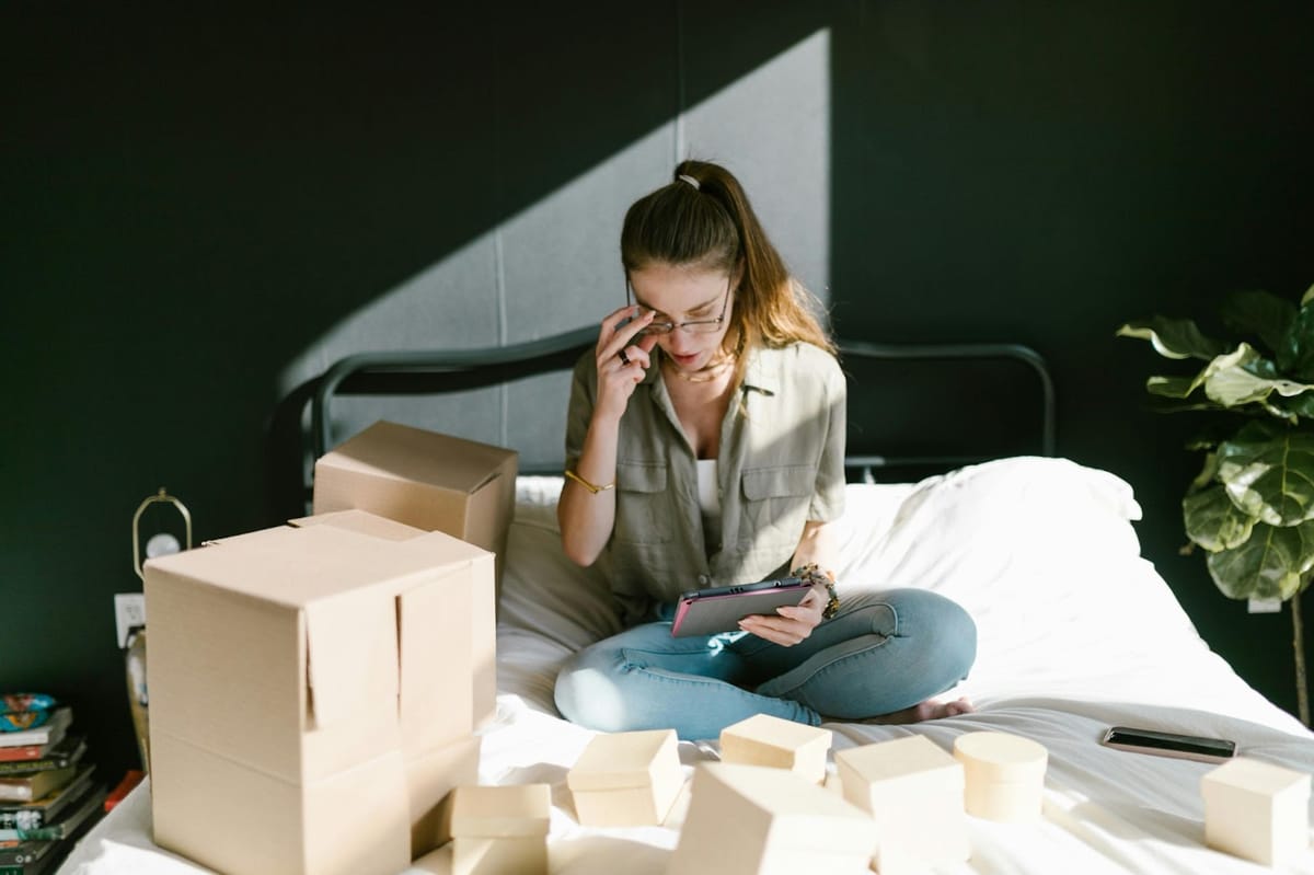 Woman with a ponytail sits on a bed, holding a tablet and adjusting her glasses, surrounded by open cardboard boxes in bright light.