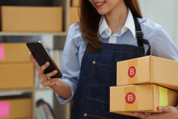 Empleada revisando el inventario en su smartphone mientras sostiene cajas de cartón con una mano