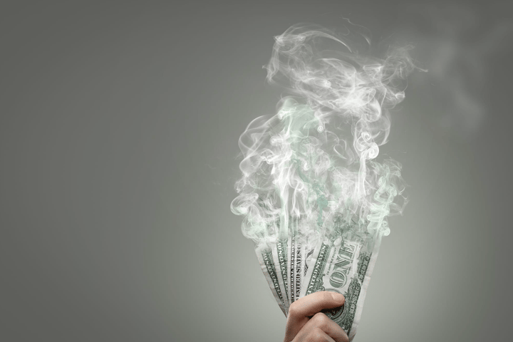 Una mano sosteniendo varios billetes de dólar estadounidense disolviéndose en el aire, asemejándose al humo.