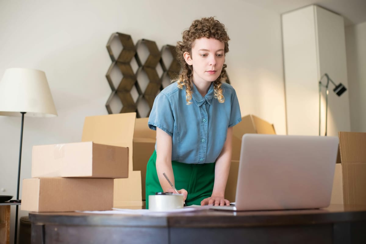 Young woman using a laptop at a desk, surrounded by cardboard boxes, possibly managing inventory or processing orders.