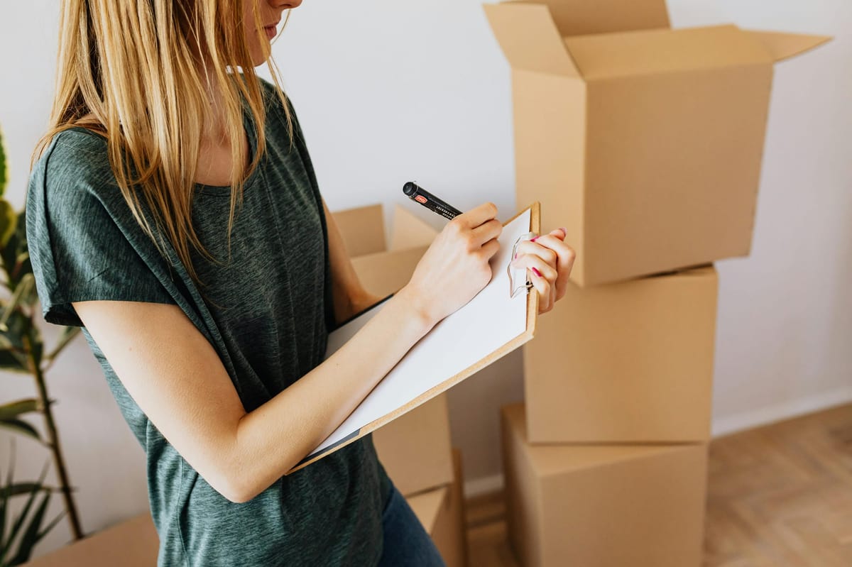 Close-up of a woman writing on a clipboard, surrounded by stacked cardboard boxes in a home or small business setting.