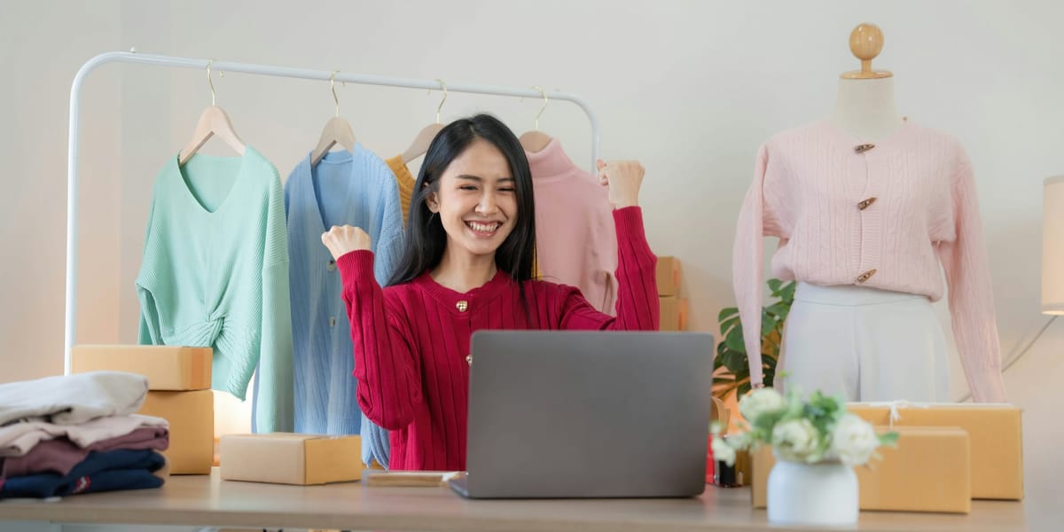 A smiling woman celebrates in front of her laptop, surrounded by clothes and boxes in a home-based online shop.