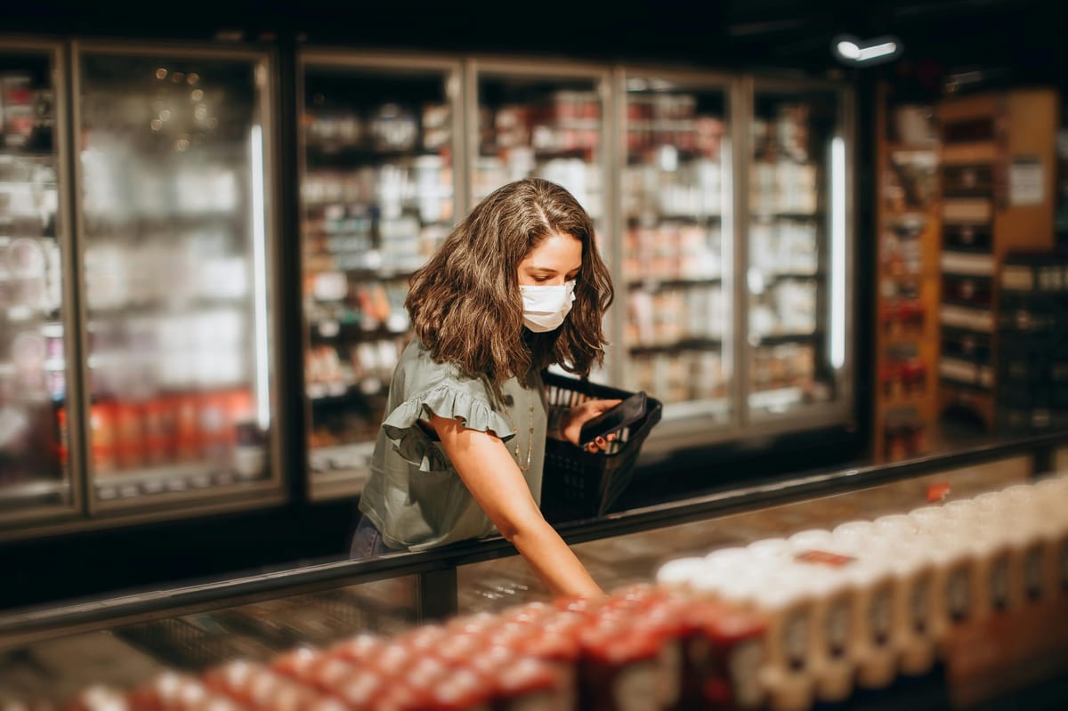 Masked woman shopping in a refrigerated section of a grocery store, selecting perishable food items.