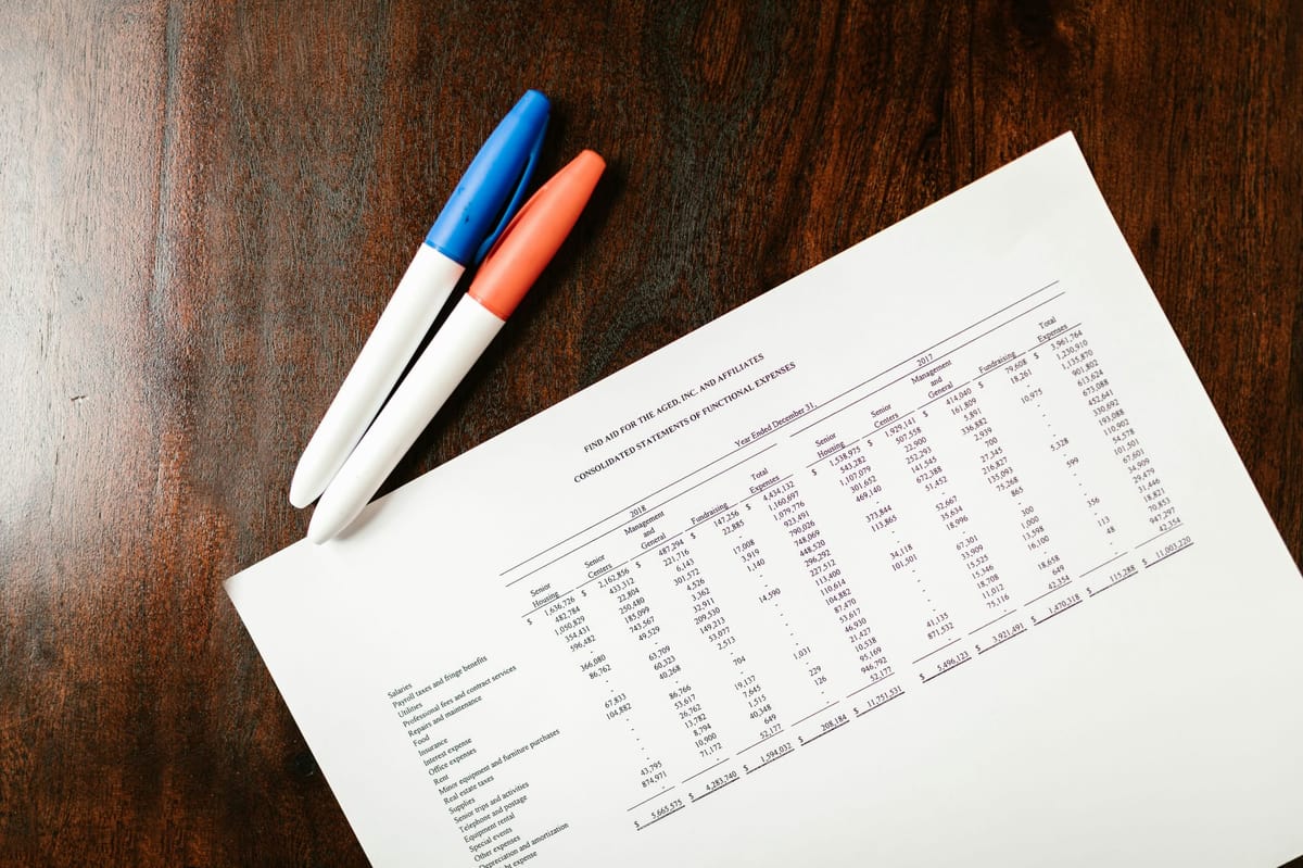 Printed financial statement with two pens on a wooden desk showing expense data.