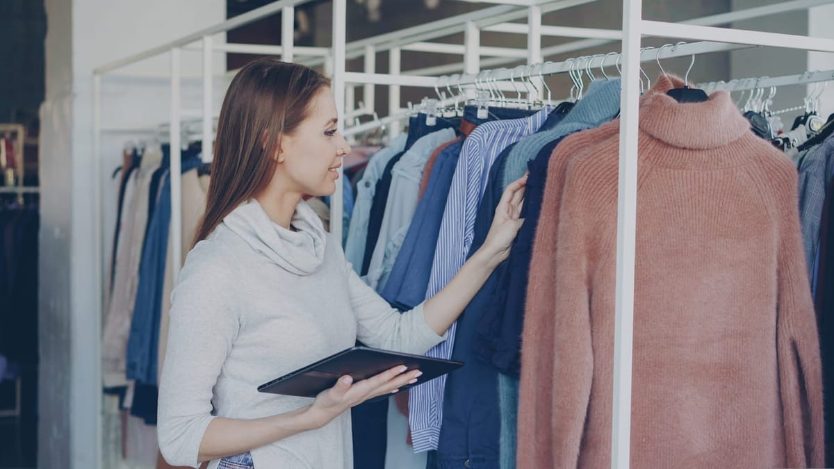 Woman Organizing a Rack of Clothing with a Tablet in One Hand