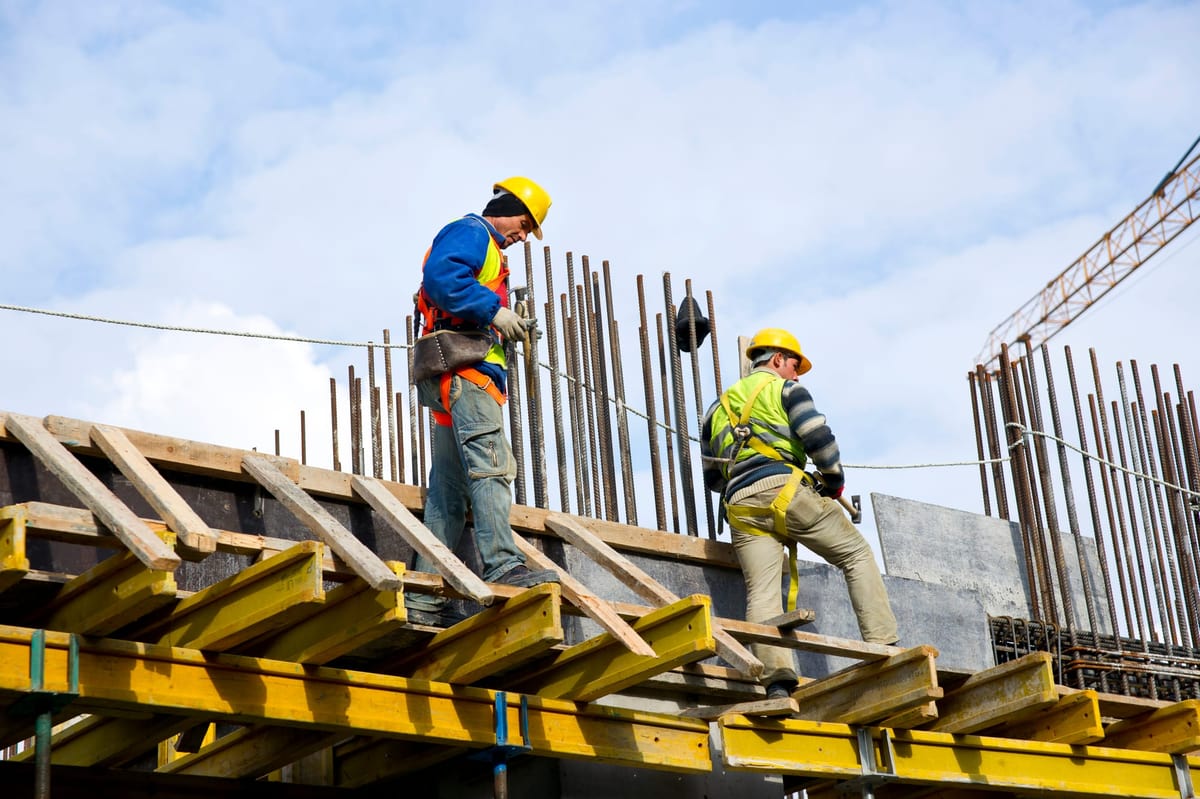 Construction workers wearing safety helmets and harnesses working on a concrete structure with exposed steel rebar.