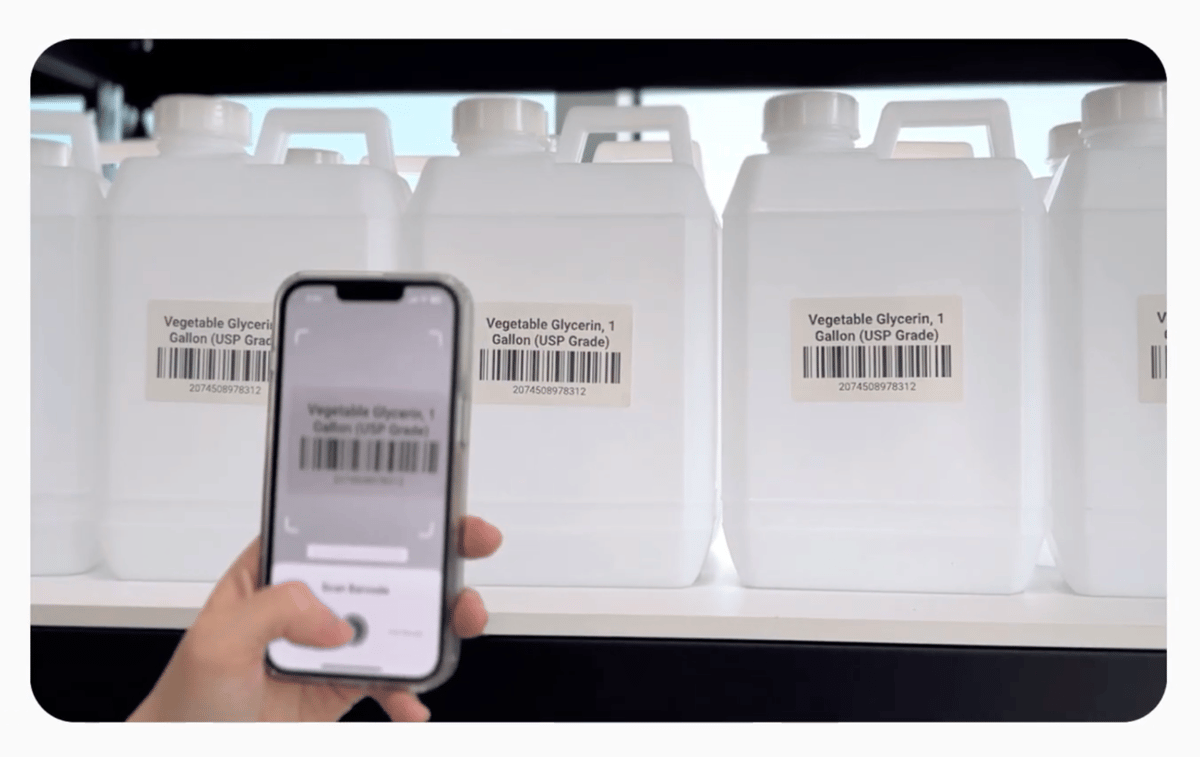 A hand scans a barcode label on a gallon jug of vegetable glycerin using the BoxHero app on a phone