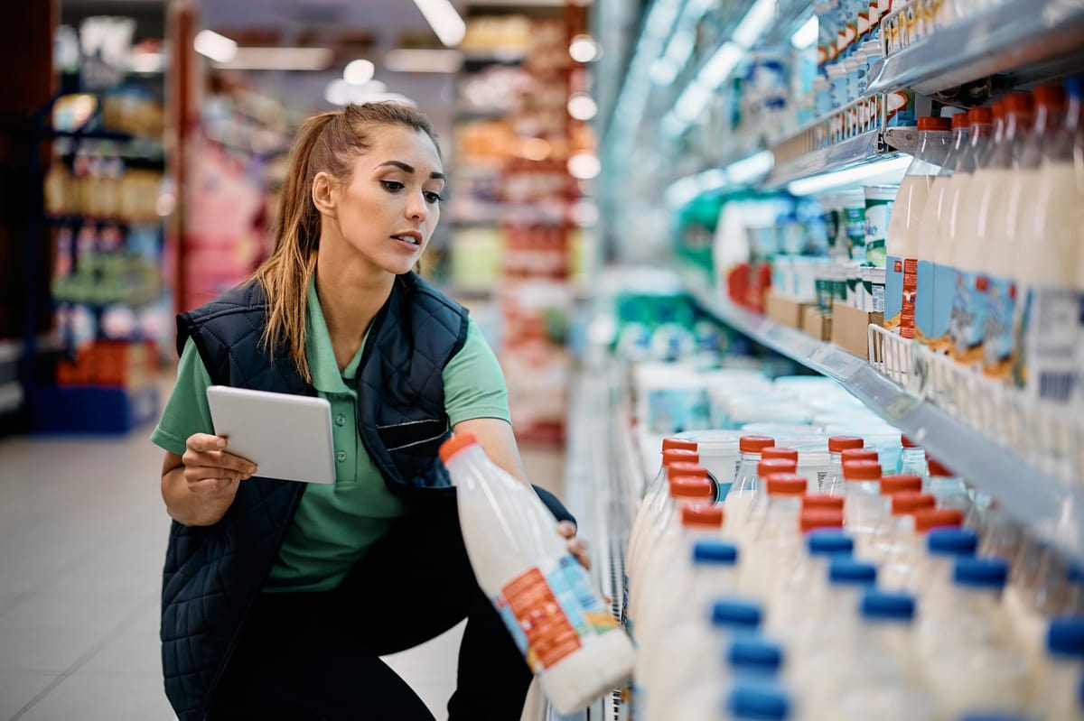 Grocery store worker holding a tablet and checking dated milk bottles on a refrigerated shelf while rotating stock.
