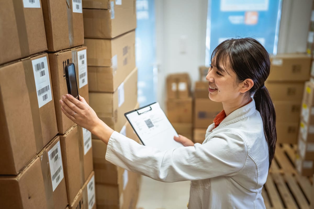 A Japanese woman scanning boxes with a phone, clipboard in hand, in a pharmaceutical warehouse