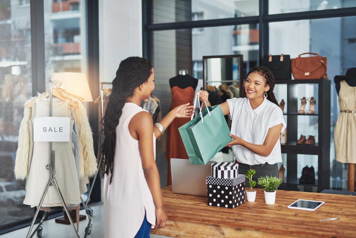 A smiling shop assistant hands a green shopping bag to a customer at the counter of a clothing boutique, with a "SALE" sign on a nearby rack.