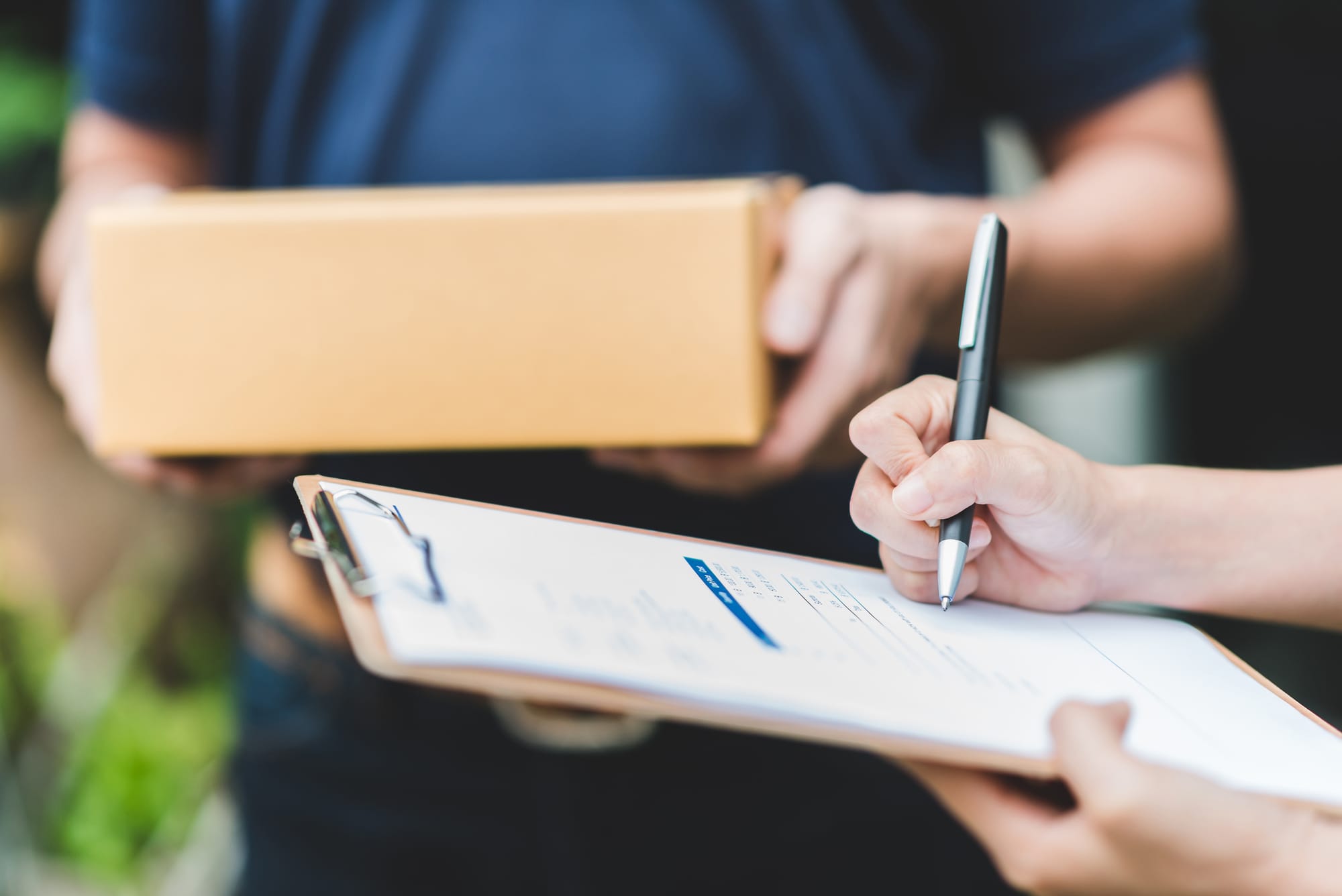 A delivery worker hands over a cardboard box while a person signs a paper delivery form on a clipboard.