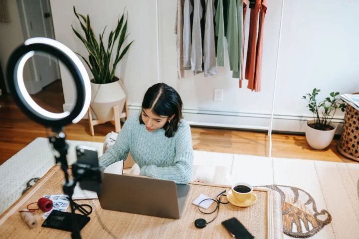 Person working at a low table with a laptop and ring light in a home studio; clothing rack and plants behind.