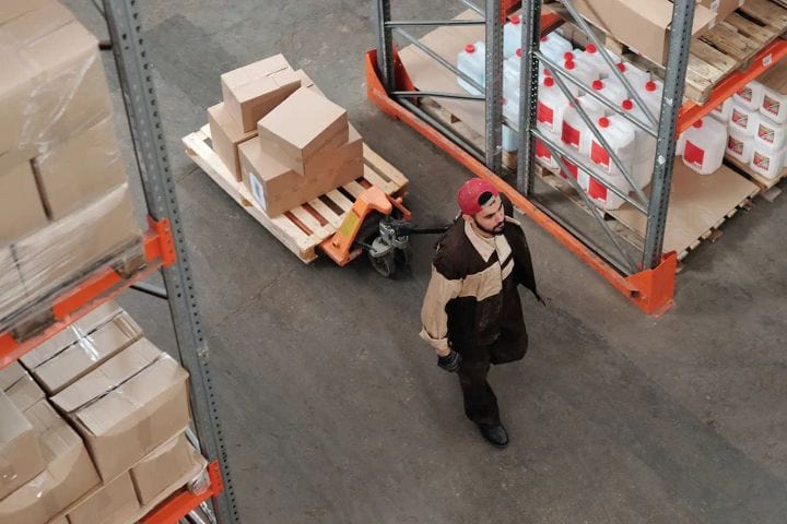 Warehouse worker pulling a pallet jack loaded with cardboard boxes down an aisle between metal racks.