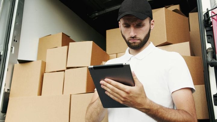 A person wearing a black cap and white polo shirt stands in a loading area, looking at a tablet while surrounded by stacked cardboard boxes.