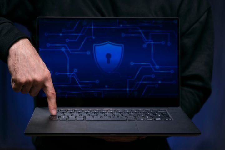 Person pointing to a laptop keyboard while holding a device showing a digital lock icon and cybersecurity protection interface