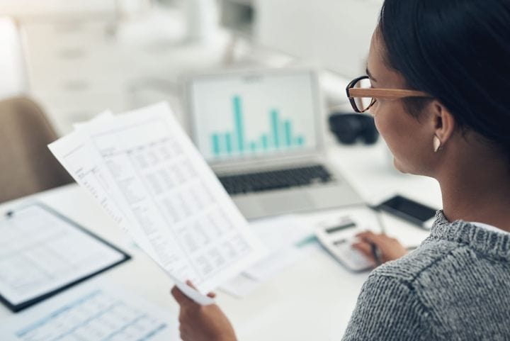 Accountant reviewing printed inventory valuation reports at a desk, comparing the figures against a bar chart on a laptop with a calculator nearby.