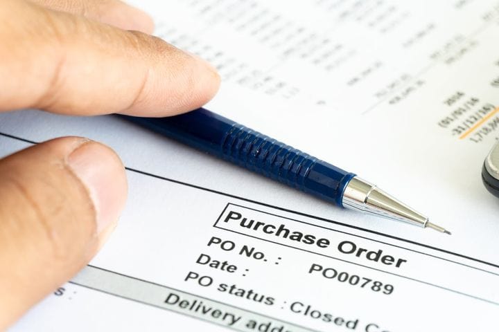 Close-up of a printed purchase order form with PO number and status, reviewed by hand with a pen