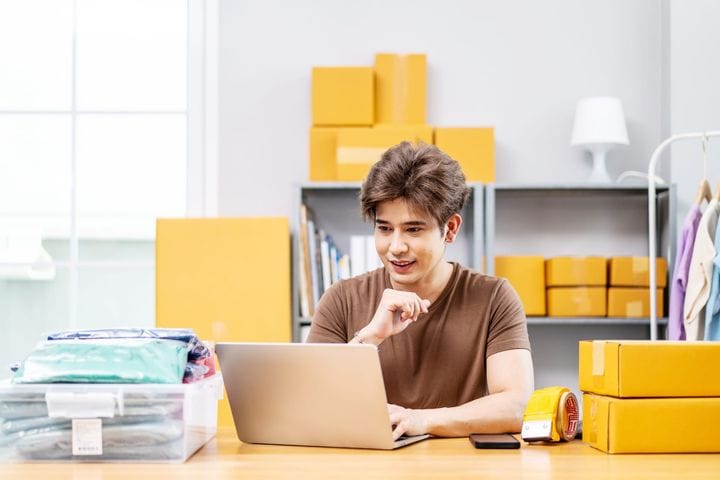 Small business owner working in home office using computer laptop for online sales and inventory management, surrounded by packaged boxes