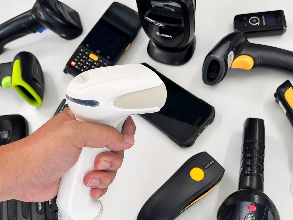 A hand holds a white Honeywell barcode scanner in the foreground, with more than ten other handheld scanners and rugged mobile computers scattered on a white desk