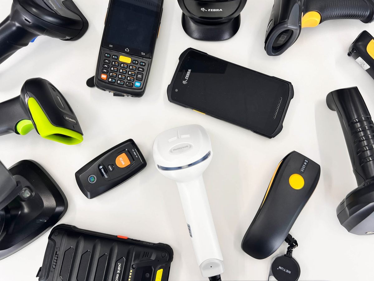 Overhead view of more than ten barcode scanners and rugged Android PDAs arranged on a white desk, including devices from Zebra, Netum, Honeywell, and other brands.